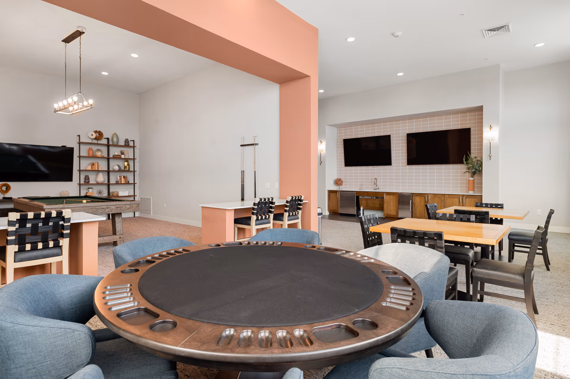Spacious communal game room with a round card table in the foreground, dining tables and chairs, two wall-mounted TVs, and a pool table in the back.