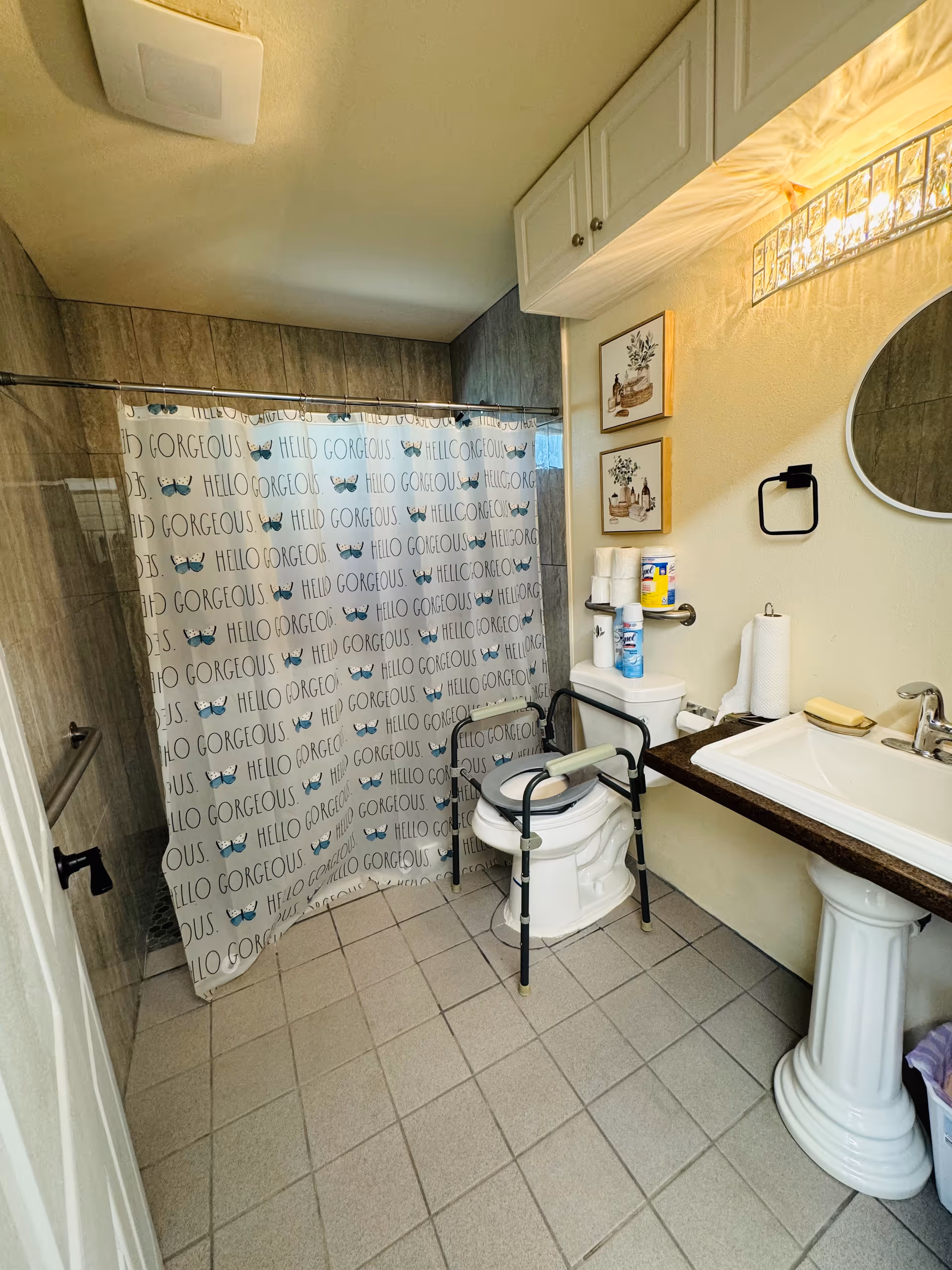 A bathroom with a tiled floor and walls, featuring a shower area with a curtain that has the words 'HELLO GORGEOUS' and butterfly designs. There is a toilet with a safety frame around it, a white pedestal sink with a dark countertop, a round mirror above the sink, and a wall-mounted light fixture. Various bathroom supplies are placed on and around the toilet, and two framed pictures hang on the wall above the toilet.