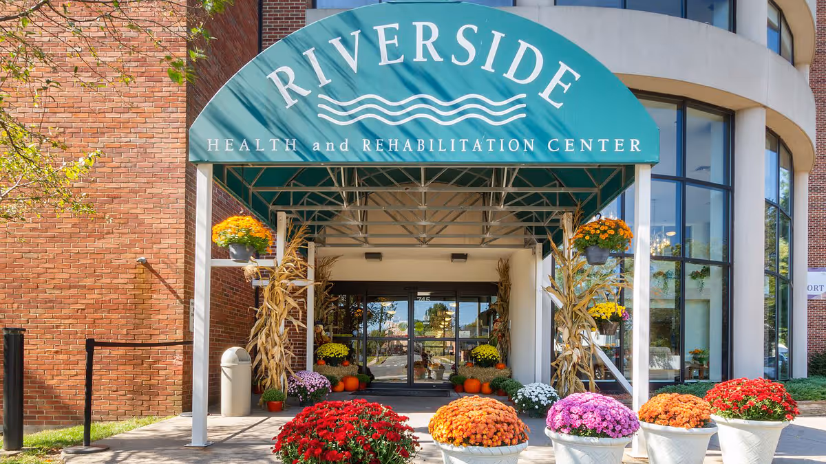 Entrance of Riverside Health and Rehabilitation Center with a green awning displaying the facility's name. The entrance is decorated with colorful potted flowers and pumpkins, with dried corn stalks on either side of the doorway. The building features large windows and brick walls.