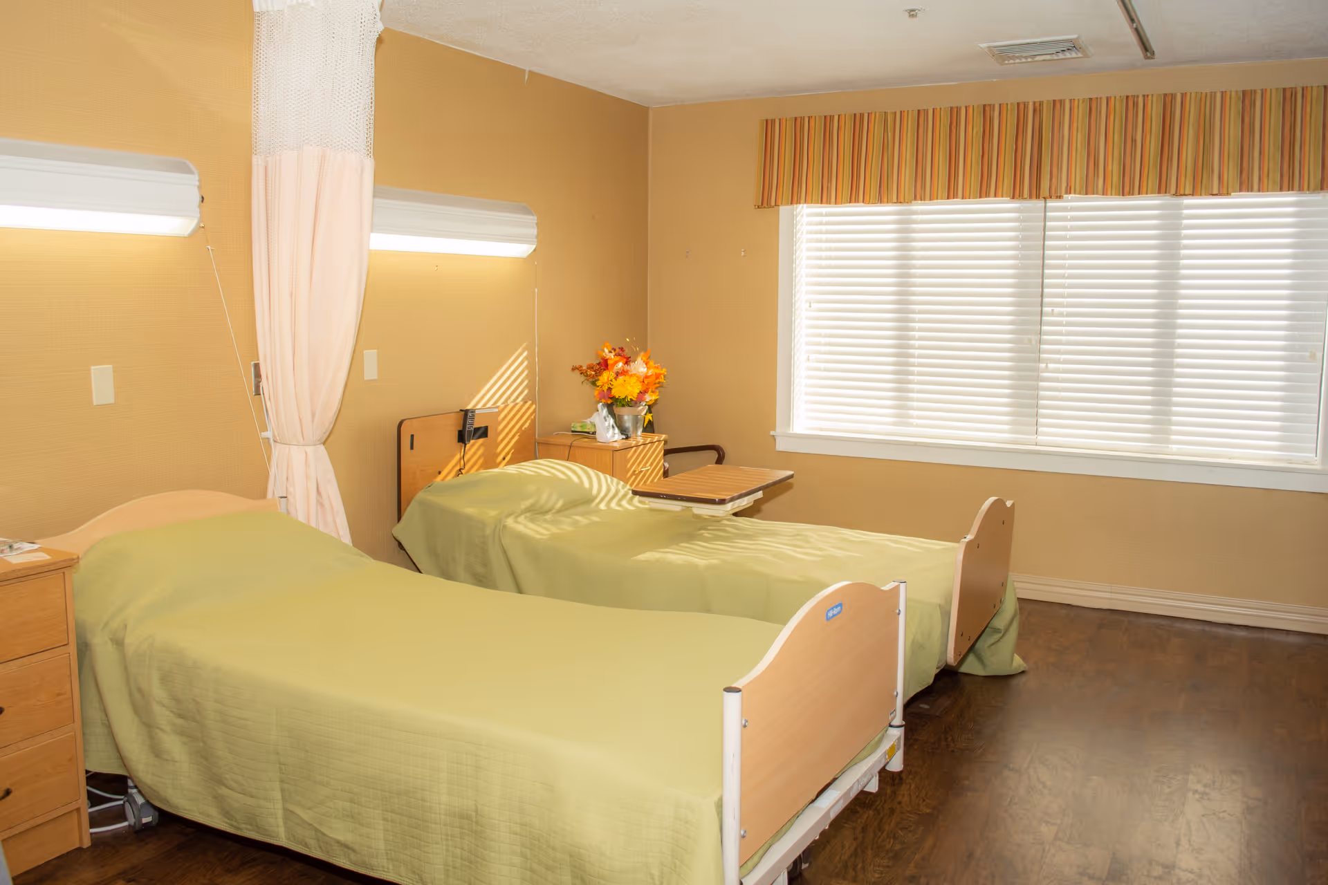 Two twin hospital-style beds with green bedspreads in a senior care room with a curtain divider, window blinds, and a small bedside table with flowers.