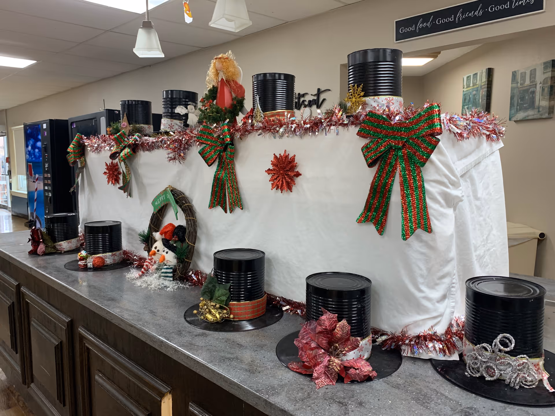 A festive holiday display on a countertop featuring black tin cans decorated as snowmen hats with various ribbons and ornaments. The display is adorned with red and green bows, tinsel garlands, and a small snowman figure. The background shows a vending machine and a sign that reads 'Good food. Good friends. Good times.'