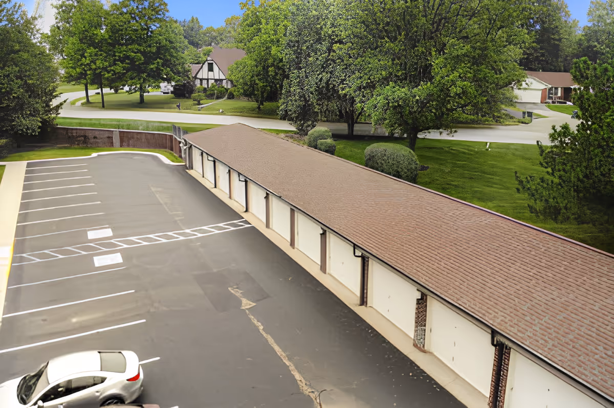A parking lot with marked parking spaces adjacent to a long building with multiple garage doors. The area is surrounded by green trees and grass, with residential houses visible in the background under a clear blue sky.
