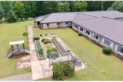 Aerial view of a single-story nursing facility building with a metal roof, surrounded by a grassy lawn and trees. There is a paved walkway leading to a small gazebo and a garden area with raised beds in the courtyard.