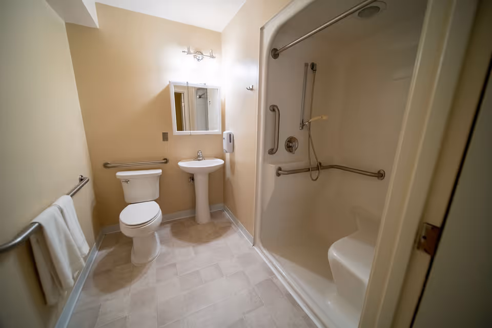 A clean and accessible bathroom featuring a toilet, a pedestal sink with a mirror above it, and a walk-in shower with grab bars and a built-in seat. The walls are painted beige, and there is a towel hanging on a rail to the left.