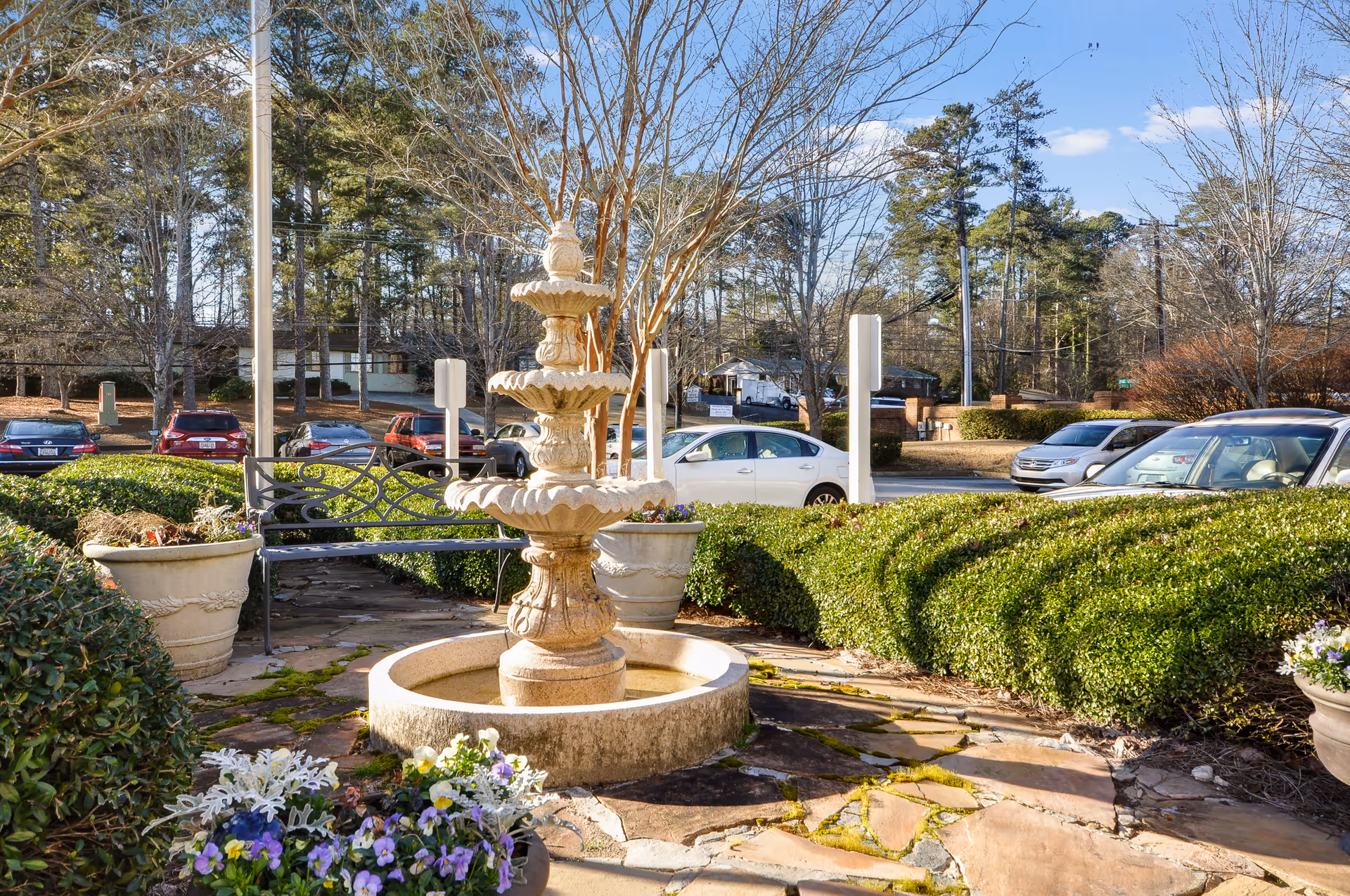 Stone tiered fountain in a landscaped courtyard with potted flowers, a bench, and parked cars visible beyond the hedges.