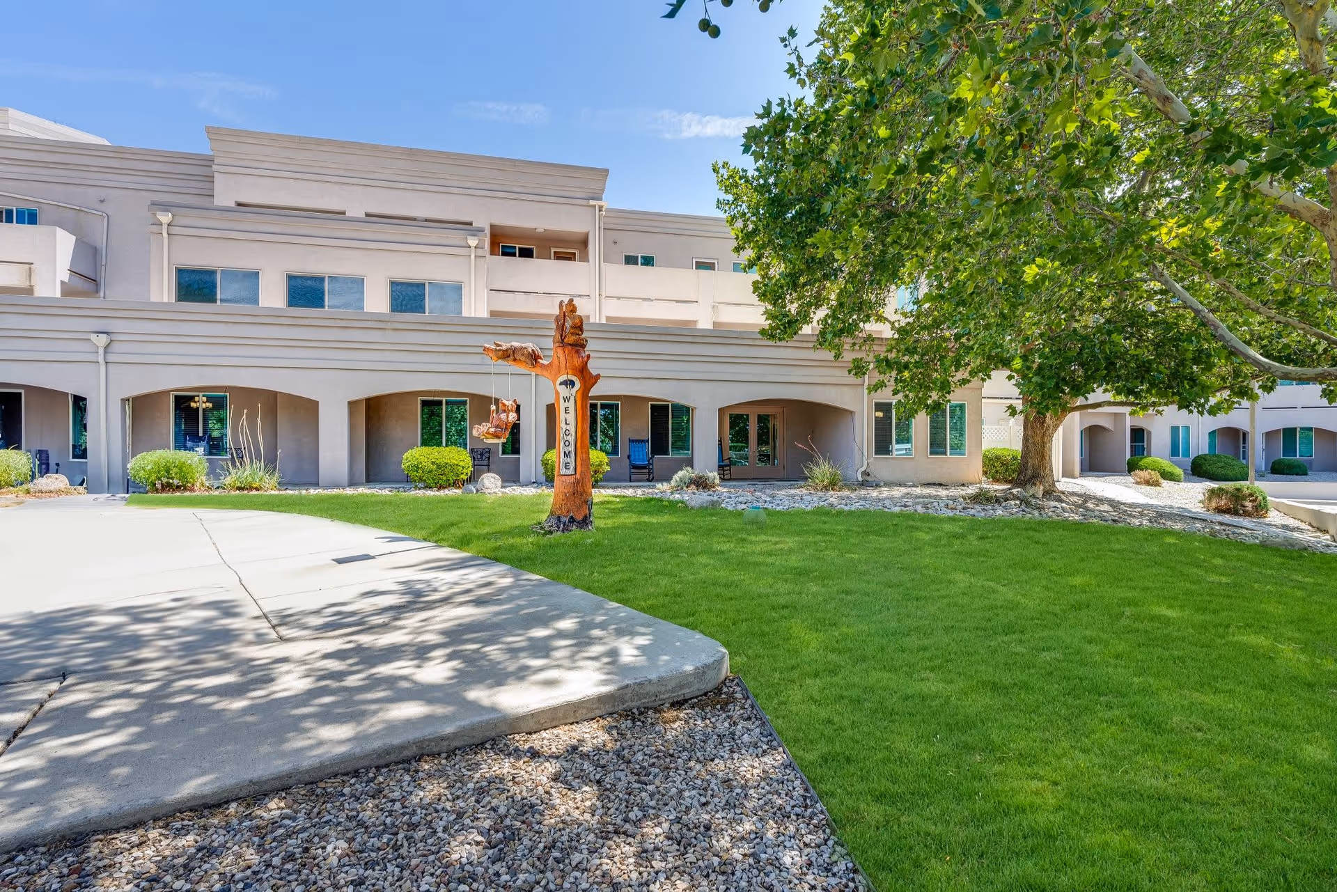 Exterior view of Bear Canyon Estates senior living facility showing a beige multi-story building with arched walkways, green lawn, a large tree, and a wooden sculpture with a welcome sign.