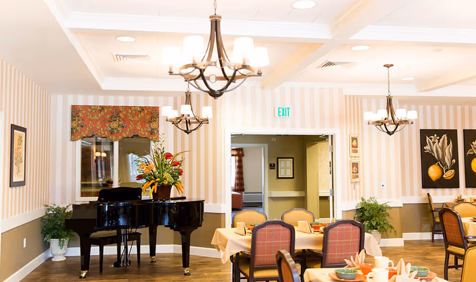 A bright dining room with tables set for a meal, featuring a black grand piano with a floral arrangement on top, striped wallpaper, framed artwork, and elegant chandeliers hanging from the ceiling.