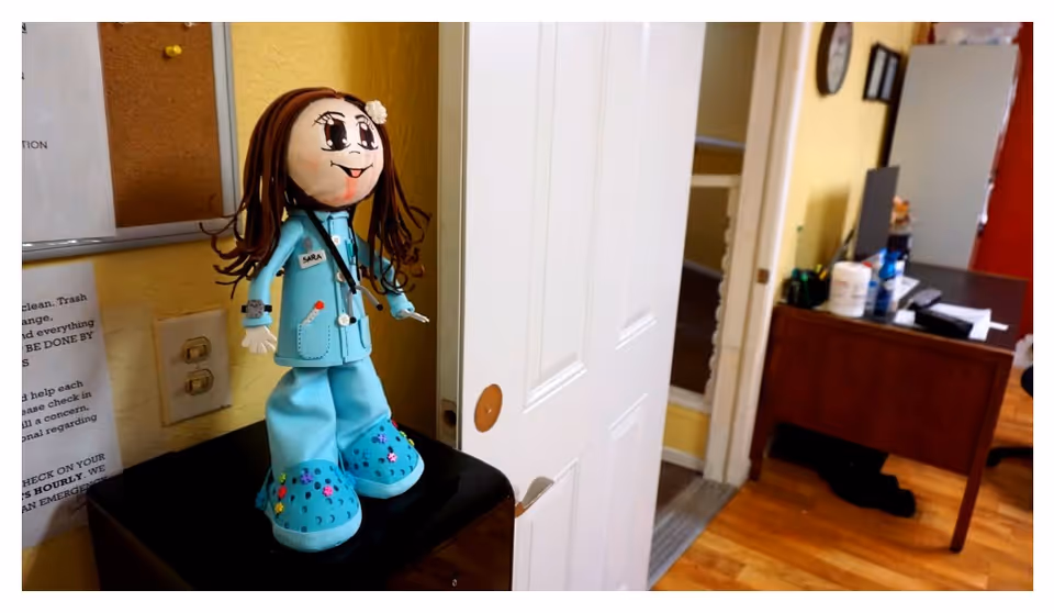 A handmade nurse doll wearing blue scrubs stands on a black surface in a reception-style interior with a partially open door and a desk in the background.