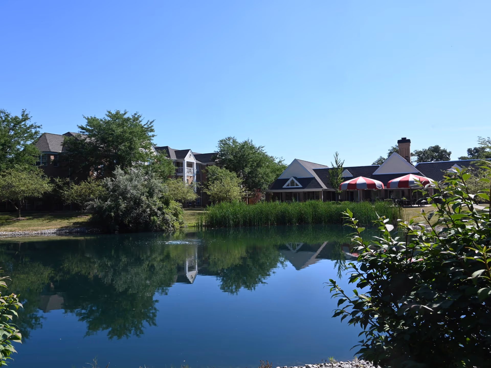 A serene outdoor scene at The Parkvue Community featuring a calm pond reflecting nearby trees and buildings. The buildings include a multi-story residential structure and a single-story building with red and white striped umbrellas on the patio. The sky is clear and blue.