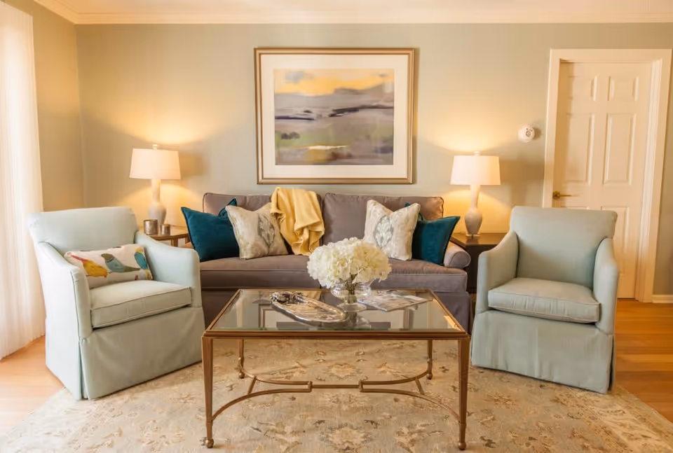 A cozy living room with a gray sofa adorned with multiple decorative pillows and a yellow throw blanket. Two light blue armchairs flank a glass coffee table with a floral centerpiece. Two table lamps on either side of the sofa provide warm lighting, and a framed abstract painting hangs on the wall above the sofa.