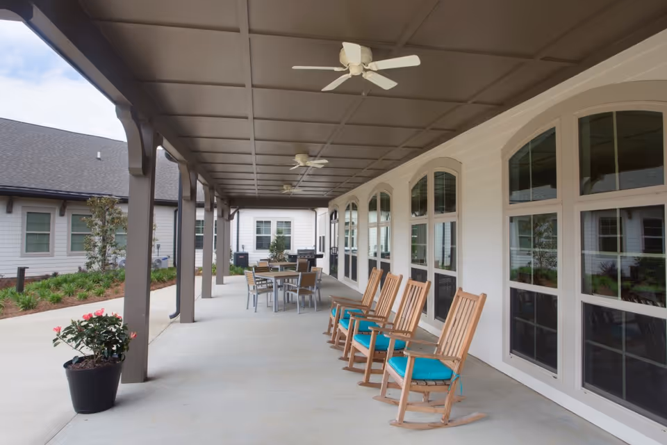 Covered outdoor patio area at The Goldton at Adelaide with wooden rocking chairs with blue cushions lined up along the wall, ceiling fans overhead, a table with chairs, and potted plants. The patio overlooks a landscaped garden area.