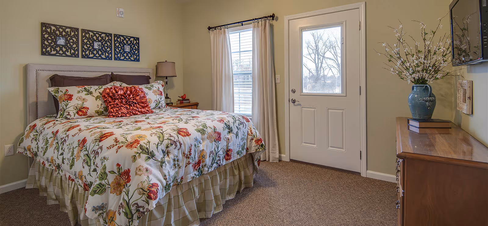 A cozy bedroom in a senior living facility featuring a bed with floral bedding and decorative pillows, a bedside table with a lamp, a window with curtains, a door with a window, a wooden dresser with a vase of flowers, and a wall-mounted television.
