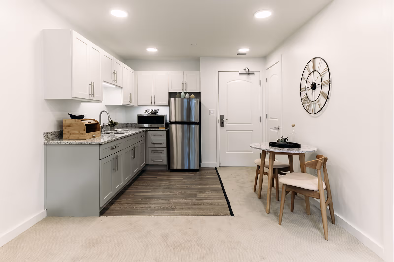 A modern kitchen and dining area in a senior living facility. The kitchen features white upper cabinets and light gray lower cabinets with silver handles, a granite countertop, a stainless steel refrigerator, a microwave, and a sink. The floor in the kitchen area is wood, transitioning to carpet in the dining area. The dining area has a small round table with a marble top and two wooden chairs with light cushions. A large decorative wall clock is mounted on the white wall above the dining table. The entrance door is visible in the background.
