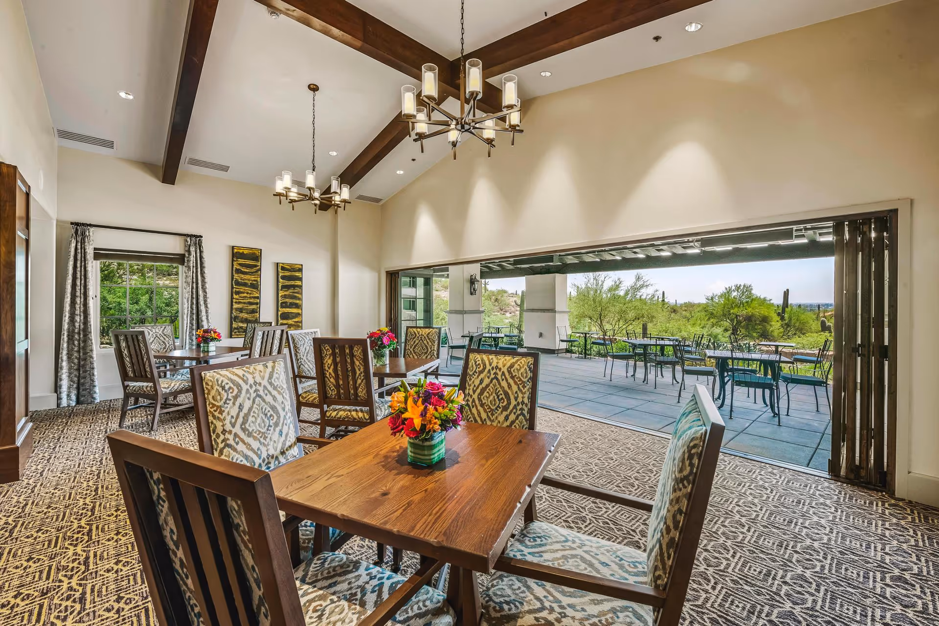 A bright dining room with wooden tables and patterned upholstered chairs, decorated with small flower arrangements. The room features high ceilings with exposed wooden beams and modern chandeliers. Large folding glass doors open to an outdoor patio area with additional tables and chairs, overlooking a scenic desert landscape with cacti and greenery.