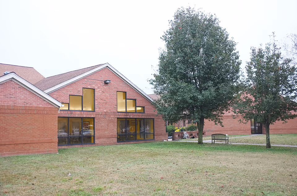 Brick exterior of an assisted living building with large windows, a grassy lawn, trees, and a bench.