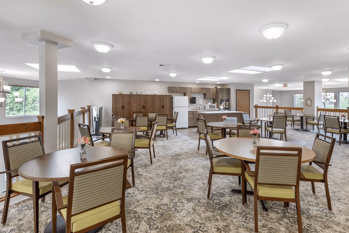 A spacious dining room in a senior living facility with multiple round tables and chairs arranged neatly. Each table has a small vase with flowers. The room features a carpeted floor, bright ceiling lights, large windows letting in natural light, and a kitchen area with cabinets, a refrigerator, microwave, and sink in the background.