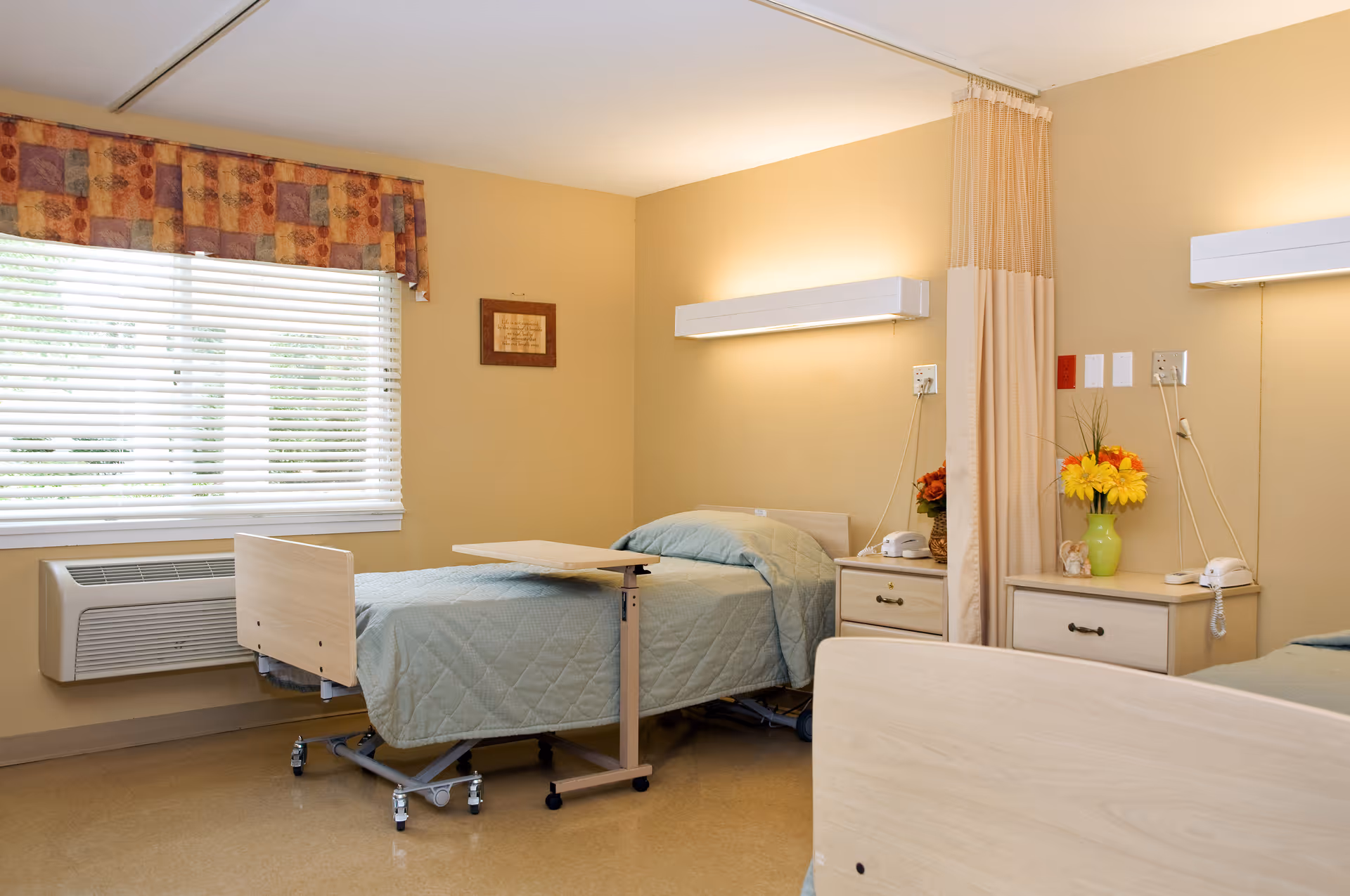 A bright senior-care bedroom with two adjustable beds, bedside tables with lamps and flowers, and a window with blinds.