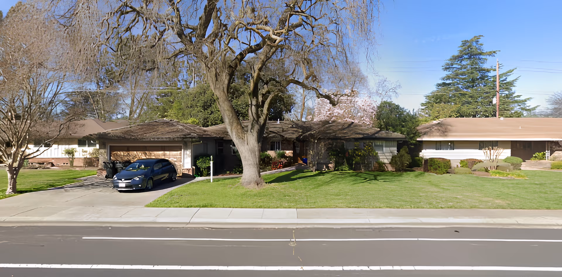 A single-story residential building with a brown roof and attached garage. There is a black car parked in the driveway. The front yard has a large tree with bare branches and well-maintained green grass. Other trees and shrubs surround the house under a clear blue sky.