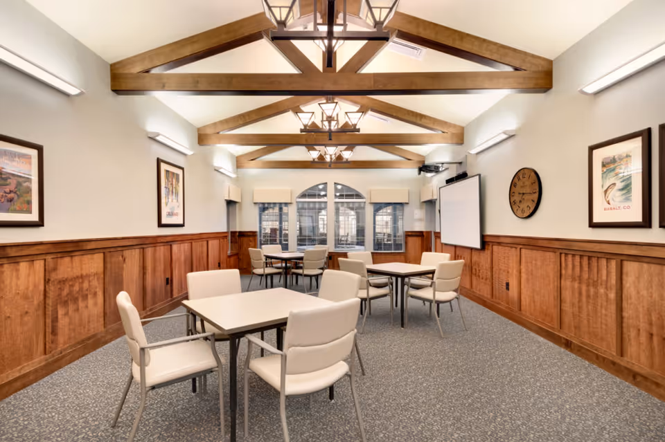 A well-lit room with wooden beams on the ceiling, several square tables each surrounded by four cushioned chairs, wood-paneled walls with framed artwork and a large clock, and a projector screen on one side. The room has carpeted flooring and windows at the far end.