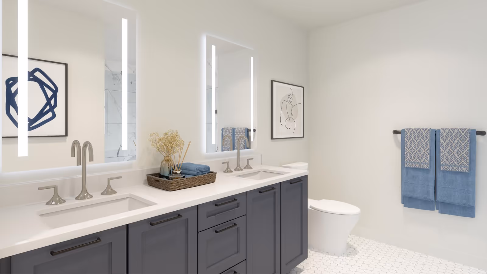 Modern bathroom with a double sink vanity featuring dark gray cabinets and white countertops. Two illuminated mirrors hang above the sinks. A decorative tray with towels and a vase with dried flowers is placed on the counter. There is a toilet next to the vanity and two blue patterned towels hanging on a towel rack on the wall. The floor has white hexagonal tiles and the walls are painted white with minimalistic framed artwork.