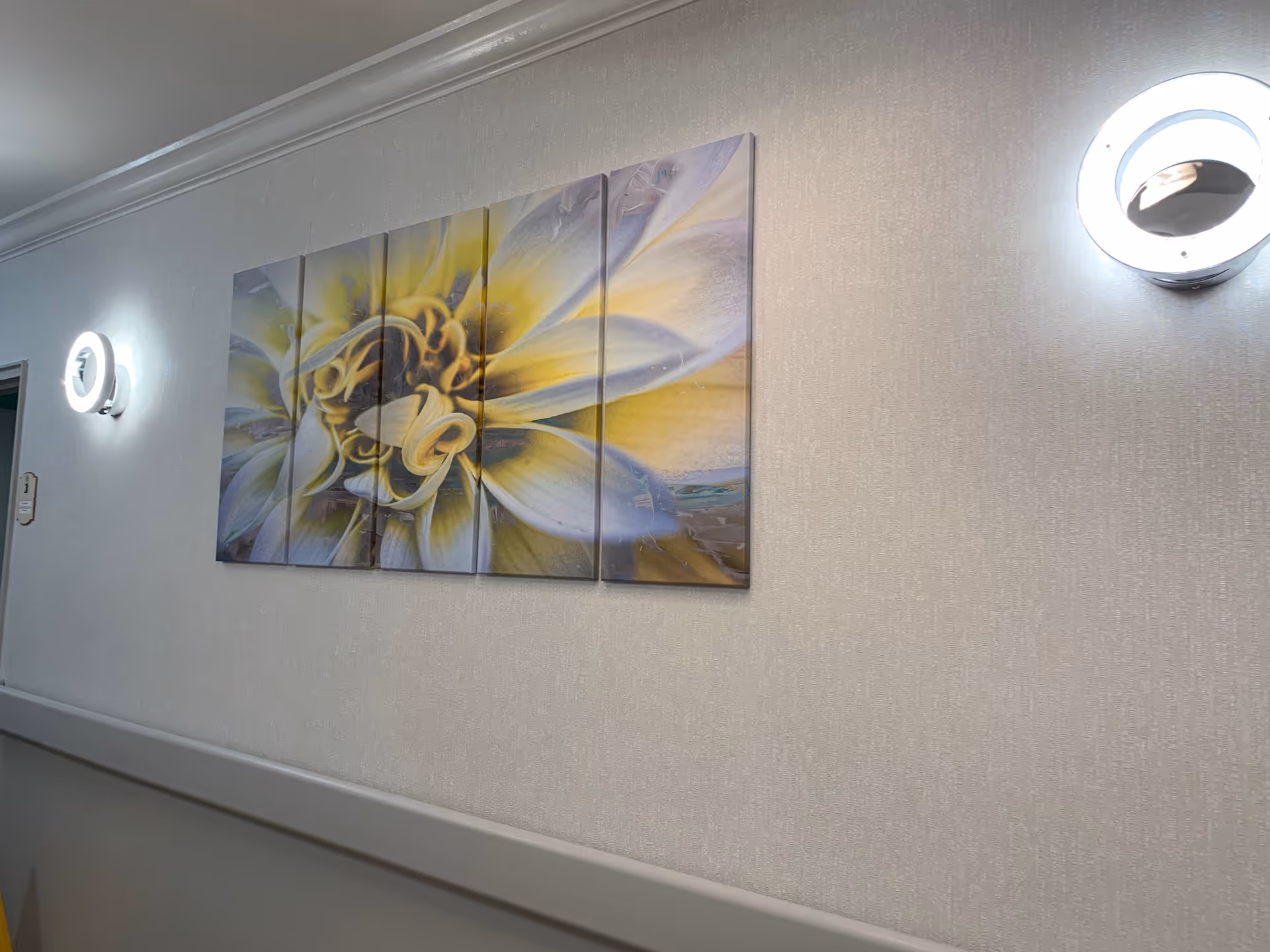 A hallway wall with a multi-panel artwork depicting a close-up of a yellow and white flower. Two circular wall lights are mounted on either side of the artwork, illuminating the textured light-colored wallpaper.