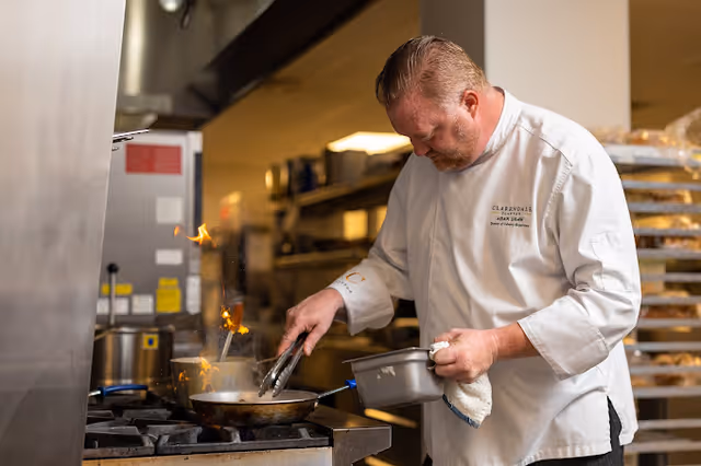 A chef in a white uniform cooking with a pan on a stove in a commercial kitchen, with flames rising from the pan and kitchen equipment and shelves in the background.