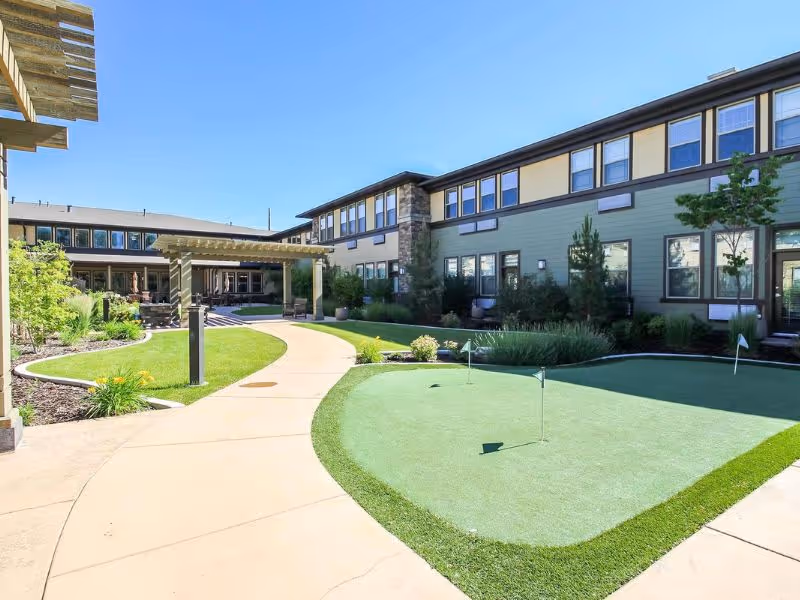 Outdoor courtyard area at Spring Gardens Senior Living Holladay featuring a putting green, curved walking paths, landscaped greenery, and a two-story building with multiple windows under a clear blue sky.