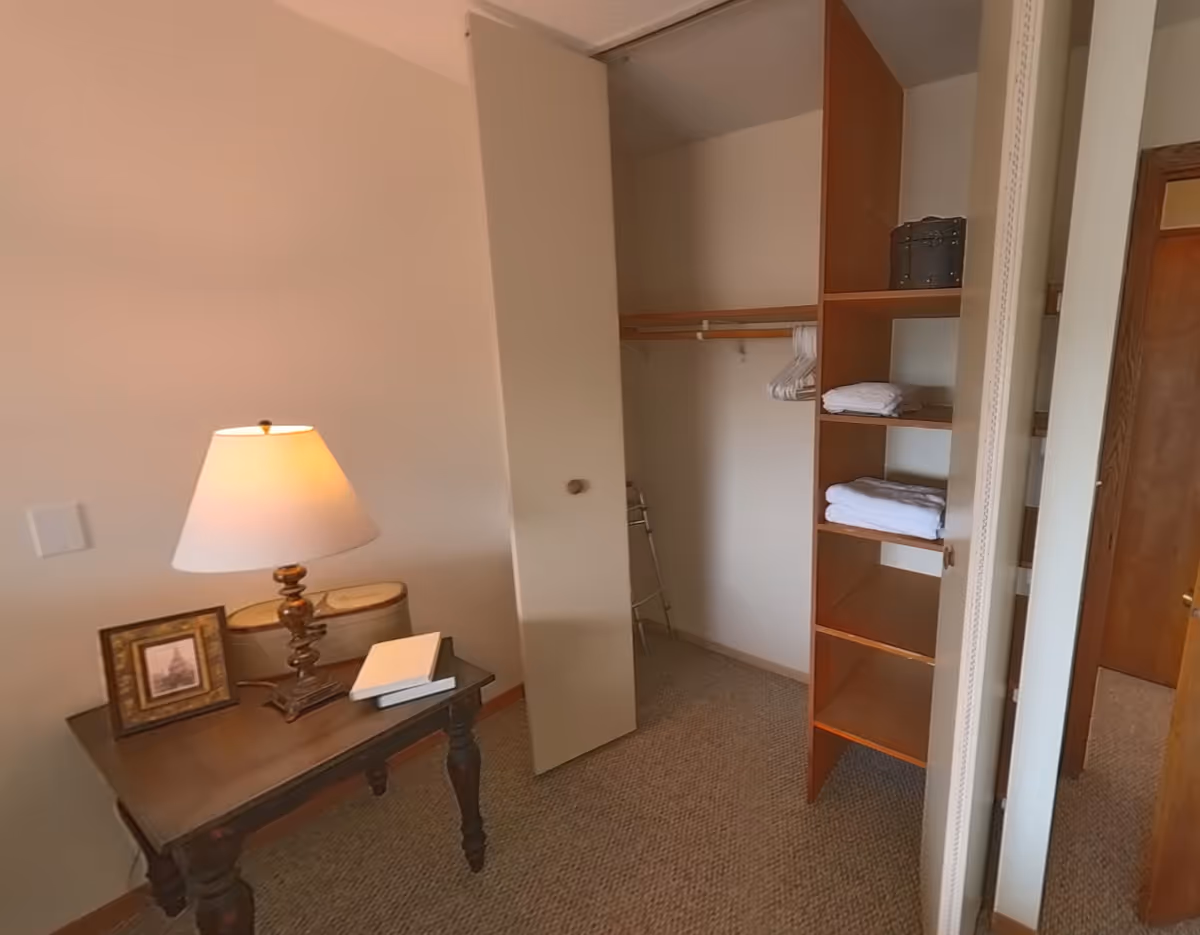 A small room with a wooden table holding a lamp, a framed picture, and two books. There is an open closet with shelves containing folded towels and hangers. A walker is partially visible inside the closet. The room has beige walls and carpeted floor.