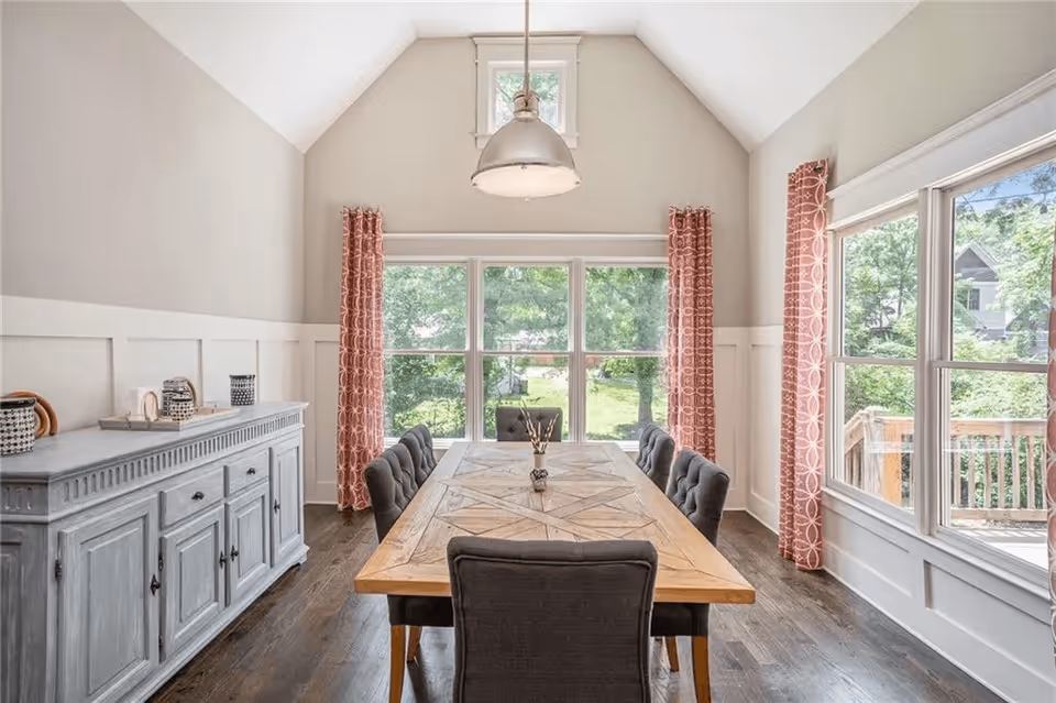 Bright dining room with a wooden table surrounded by upholstered chairs, large windows and coral patterned curtains.