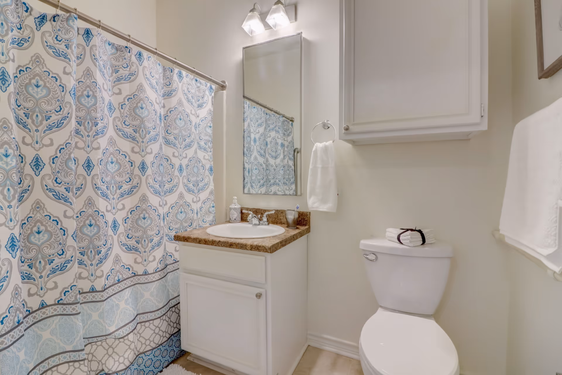 A clean bathroom featuring a white toilet with folded towels on top, a white vanity with a brown countertop and a sink, a mirror above the sink with two light fixtures, a towel ring with a white hand towel, a white cabinet mounted on the wall, and a shower with a decorative blue and gray patterned curtain.