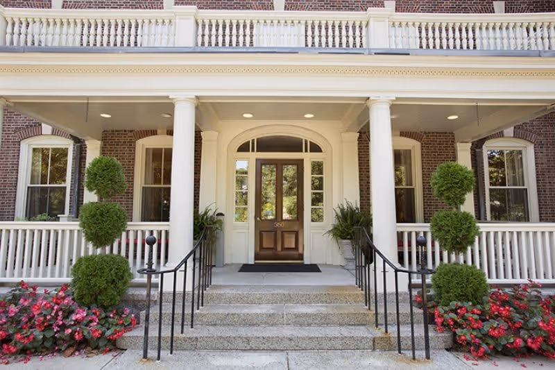 Front entrance of a brick building with white columns and railings, featuring a wooden door with glass panels, steps leading up to the door, neatly trimmed topiary plants, and red flowers on either side of the stairs.