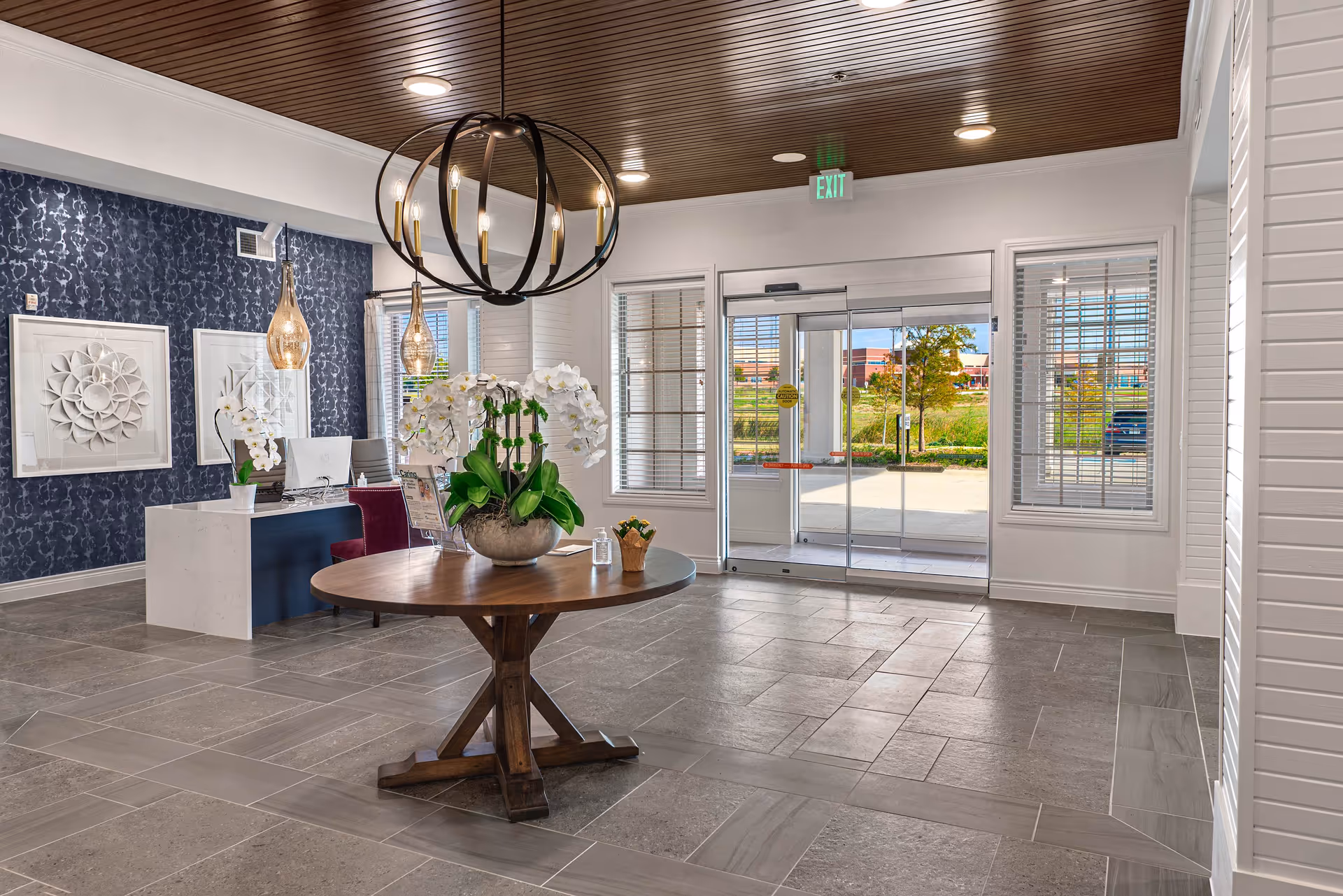 Bright modern lobby with a round wooden table topped by white orchids, a reception desk, chandelier, and glass entrance doors.