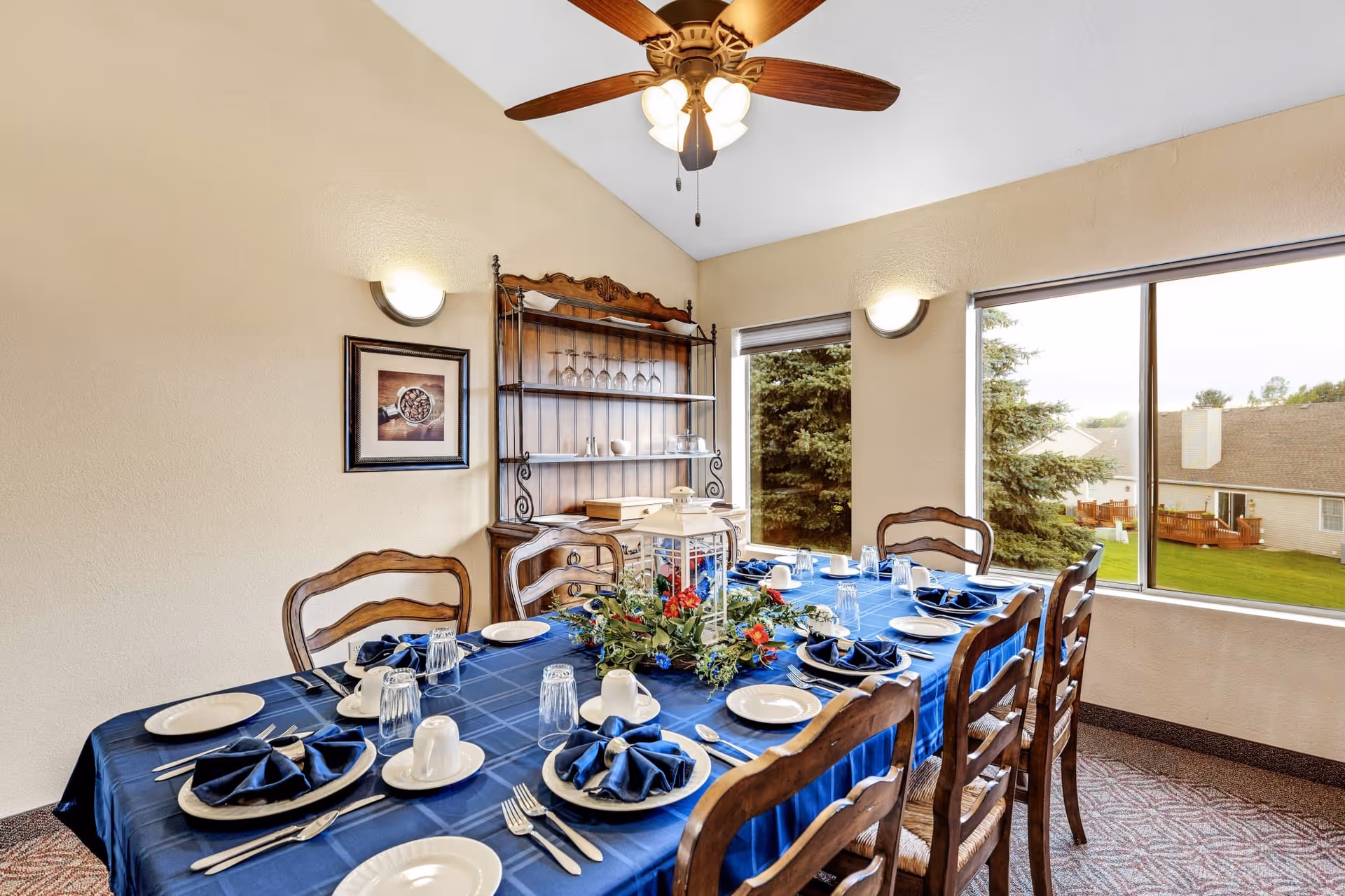 A dining room with a rectangular table covered with a blue tablecloth, set with white plates, cups, silverware, and blue folded napkins. A centerpiece with greenery and red flowers is in the middle of the table. Wooden chairs surround the table. The room has beige walls, a ceiling fan with lights, a wooden hutch with glassware, and large windows showing a green outdoor area with trees and neighboring buildings.
