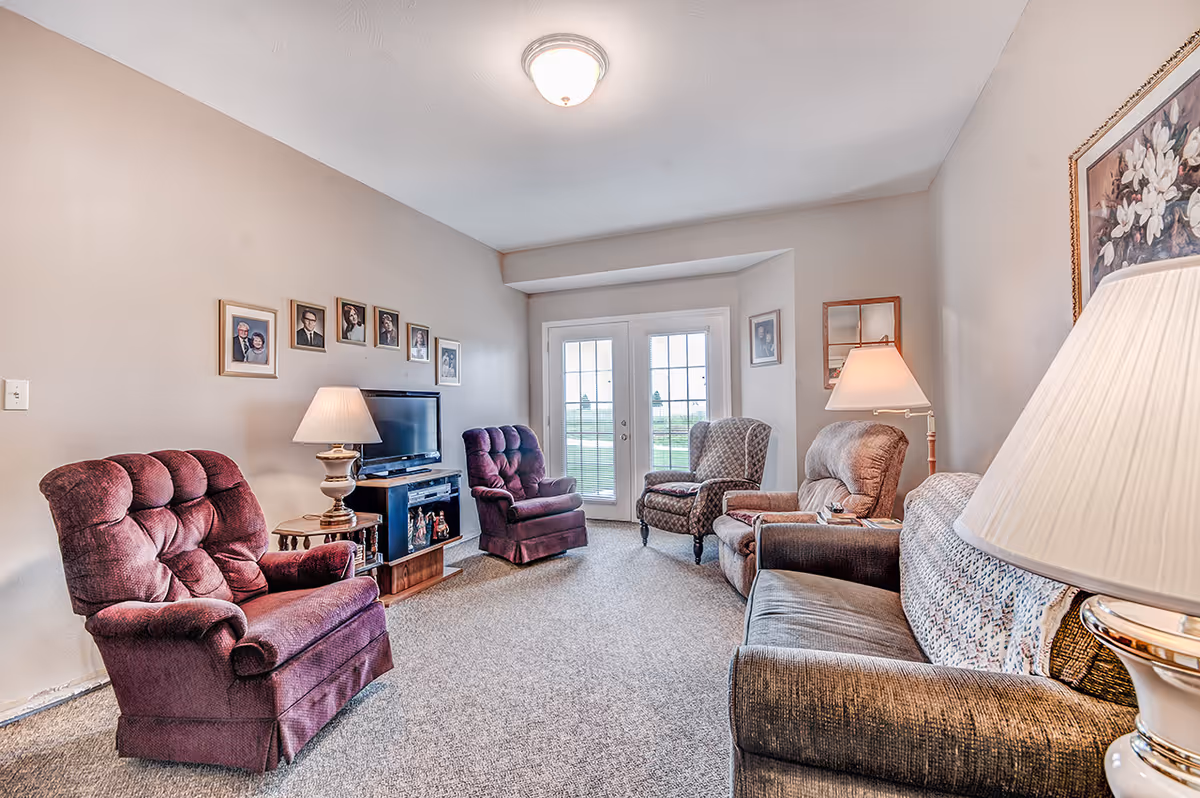 Bright living room with upholstered armchairs and a sofa arranged around a TV and lamps, with French doors at the far wall.