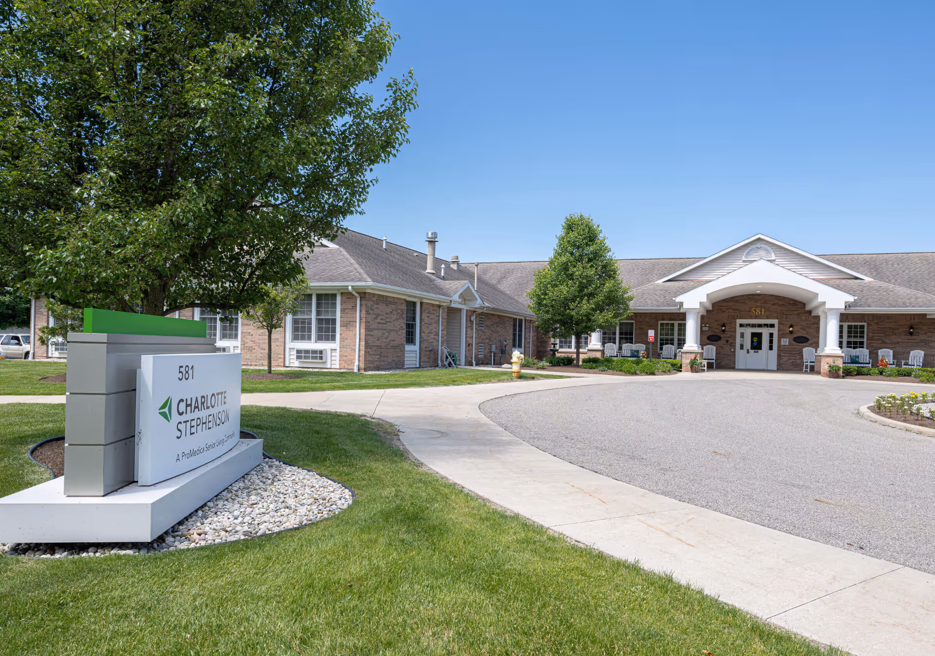 Exterior view of Charlotte Stephenson A ProMedica Senior Living Community building with a driveway, green lawn, trees, and a sign displaying the facility name and address 581.