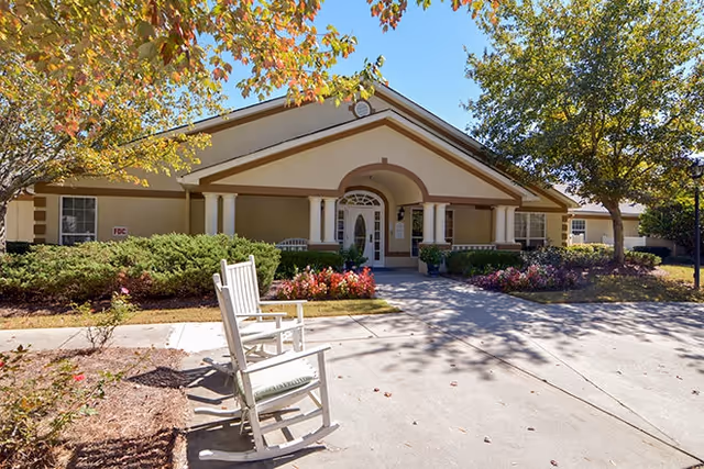 Front entrance of a single-story senior living building with a covered portico, white rocking chairs, and landscaped flower beds.