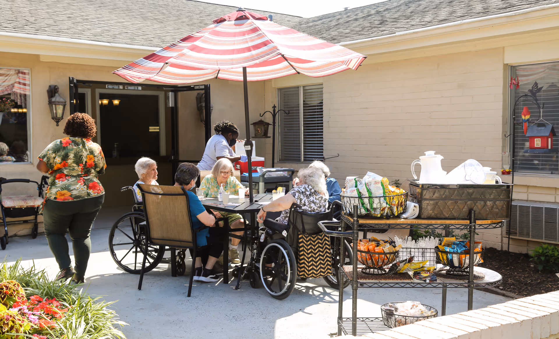 A group of elderly people, some in wheelchairs, sitting around a table under a red and white striped umbrella outside a building. A caregiver is attending to them. Nearby, there is a cart with snacks, drinks, and disposable utensils. The setting appears to be a patio area of a senior living facility.
