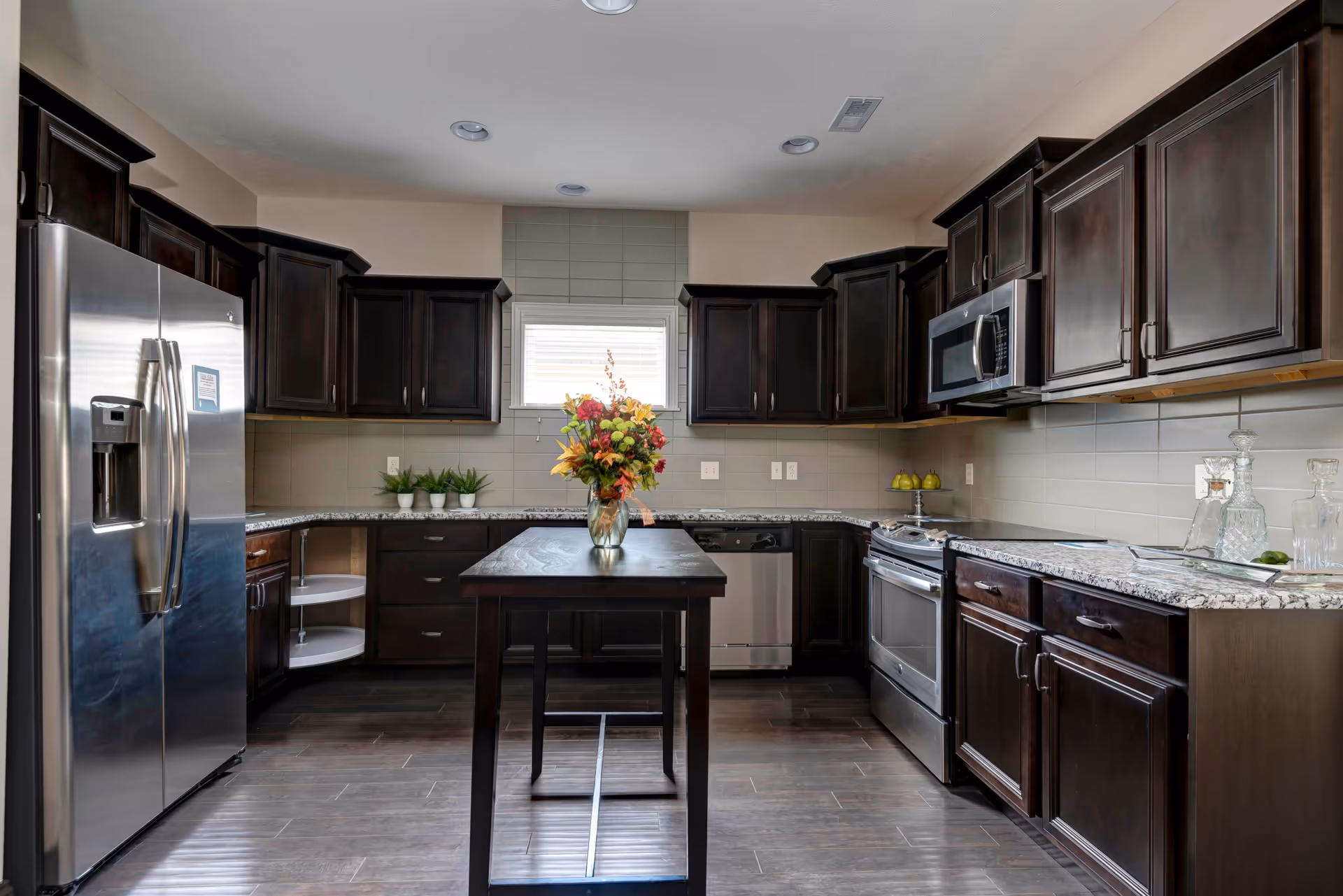Modern kitchen with dark wood cabinets, stainless steel refrigerator, oven, microwave, and dishwasher. A small island with a vase of colorful flowers is centered on the dark wood floor. A window with blinds is above the countertop, and the backsplash is tiled in a neutral color.