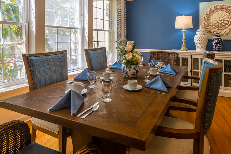 A dining room with a wooden table set for six people, featuring blue cloth napkins, white cups and saucers, water glasses, and silverware. The room has large windows letting in natural light, blue walls, a white sideboard with decorative vases and a lamp, and a floral centerpiece on the table.