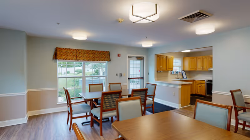 A dining room with wooden tables and upholstered chairs adjacent to an open kitchen and a windowed exterior door.