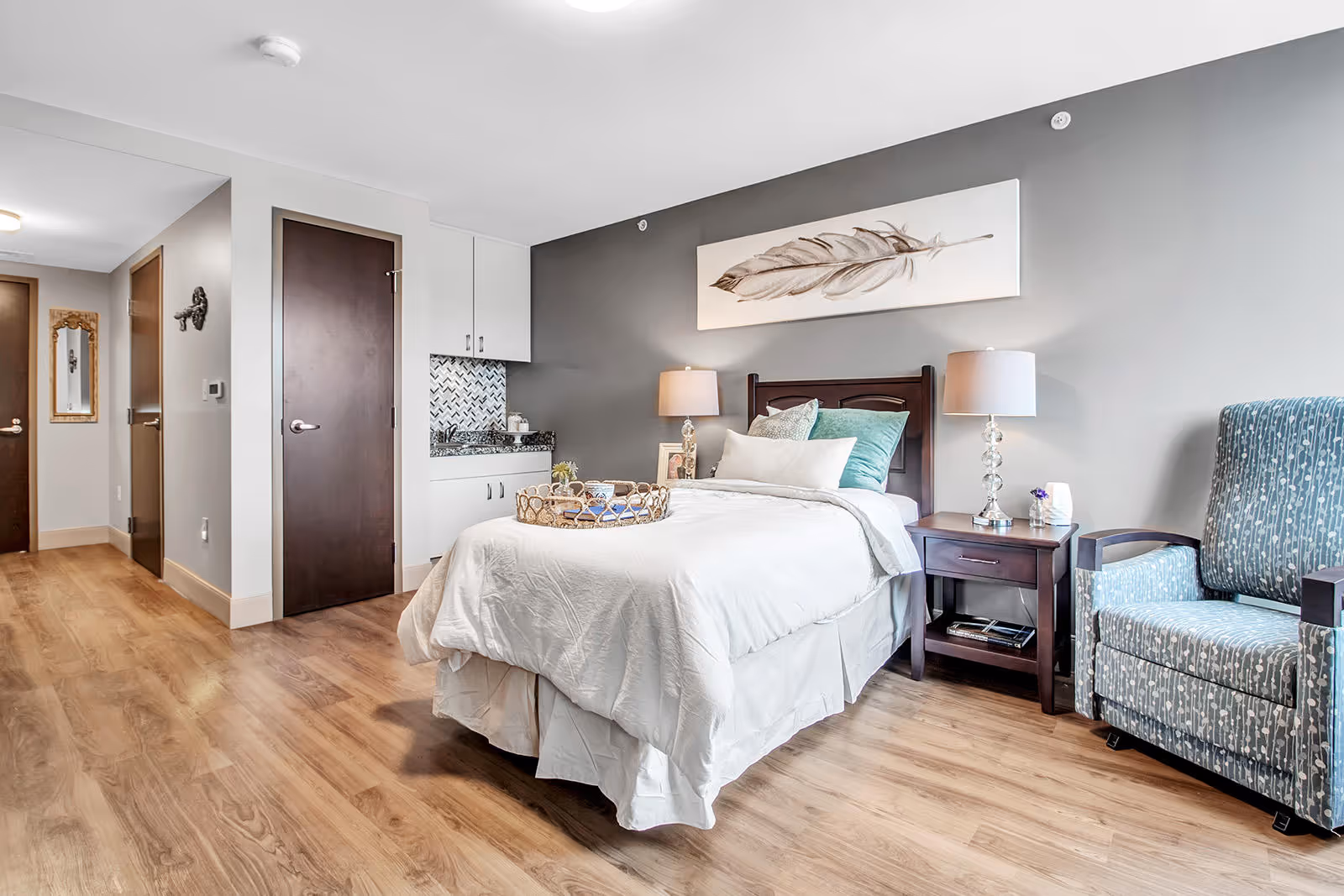 A bright and clean bedroom in an assisted living facility featuring a single bed with white bedding and multiple pillows. There is a wooden nightstand with a lamp and decorative items next to the bed. A comfortable blue patterned armchair is positioned to the right of the nightstand. The walls are painted gray and white, with a large feather artwork hanging above the bed. The room has wood flooring and a small kitchenette area with a granite countertop and white cabinets. Two closed dark wooden doors are visible in the background along with a hallway and a wall-mounted mirror.
