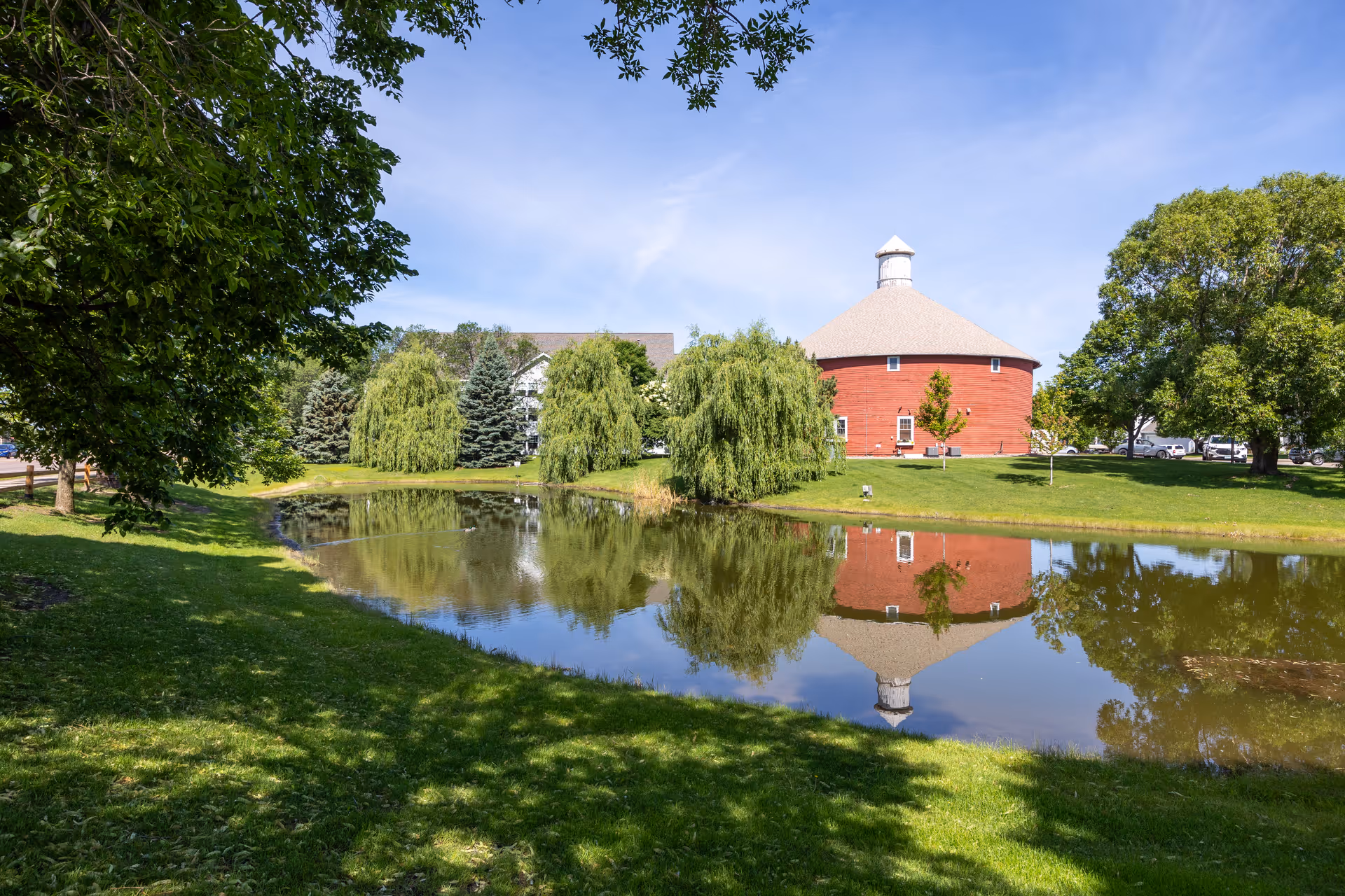 A peaceful outdoor scene at The Farmstead featuring a small pond reflecting a large round red barn-like building and surrounding trees under a clear blue sky.