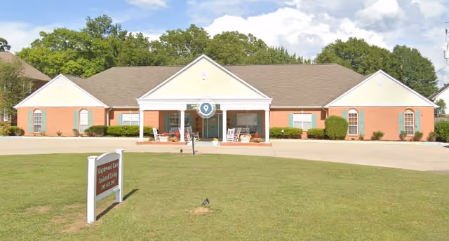 Front view of a single-story brick assisted living building with a covered entrance, rocking chairs on the porch, and a sign on the lawn.