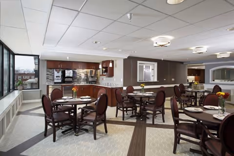 A bright dining room in a senior living facility with multiple round tables set with plates and silverware. Each table has a small vase with flowers. The room features large windows on one side, allowing natural light to fill the space. In the background, there is a kitchenette area with wooden cabinets, a coffee machine, and a countertop. The ceiling has recessed lighting and modern light fixtures.
