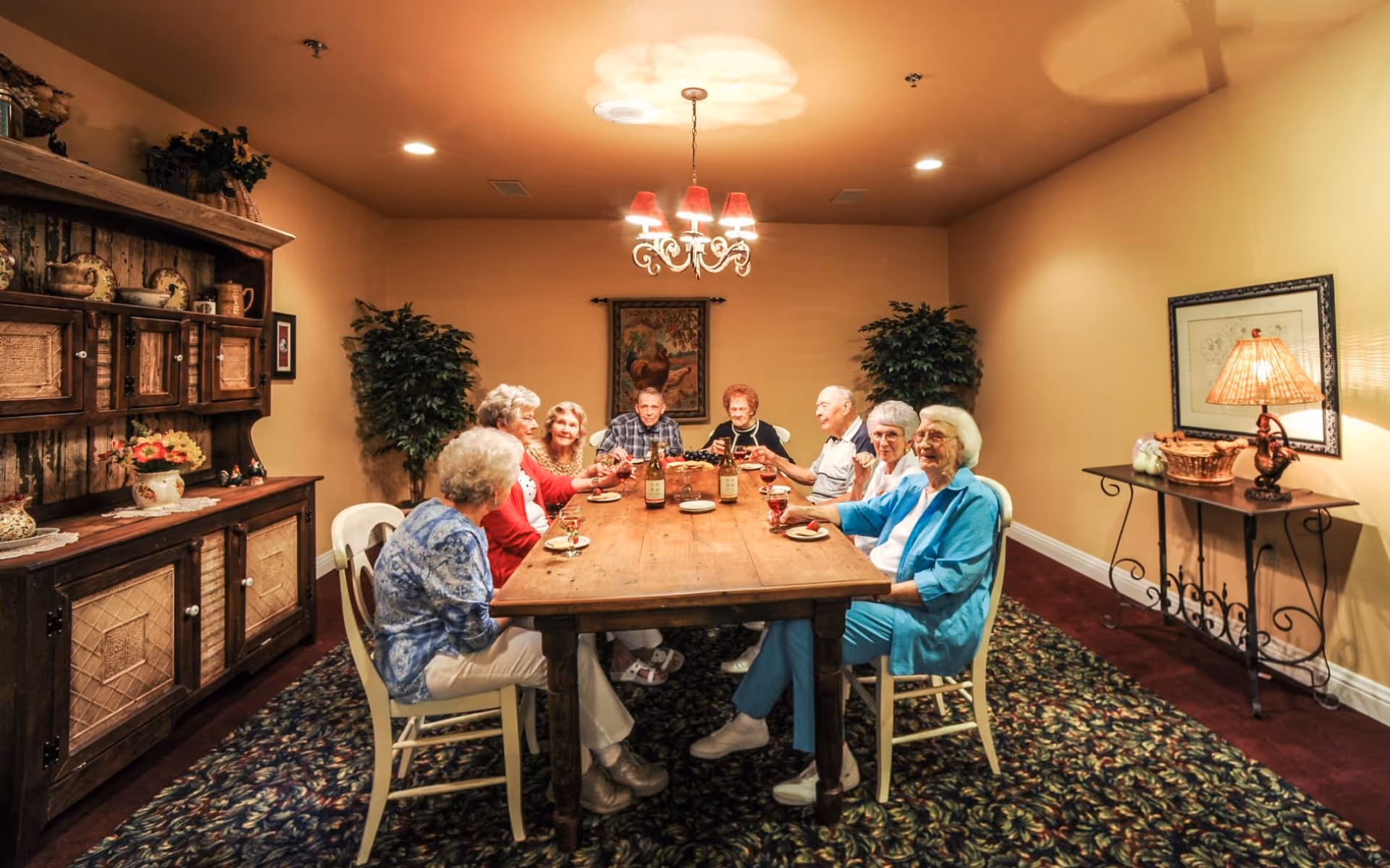 A group of elderly people sitting around a wooden dining table in a warmly lit room, enjoying drinks and snacks. The room has beige walls, a patterned carpet, a wooden cabinet with decorative items on the left, two potted plants, a chandelier with red lampshades hanging above the table, and a side table with a lamp and basket on the right.