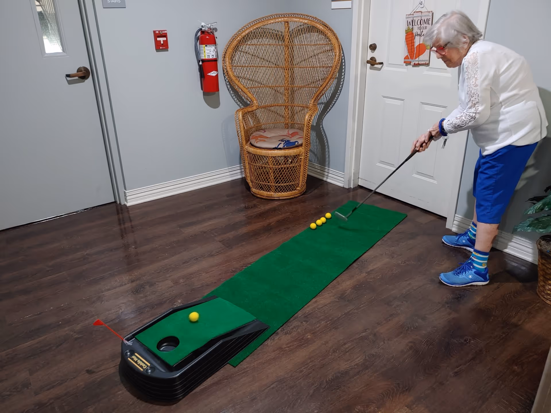 An elderly woman playing indoor mini golf on a green putting mat with four yellow golf balls lined up. She is standing on a wooden floor in a room with light gray walls, next to a wicker chair and a white door with a welcome sign.