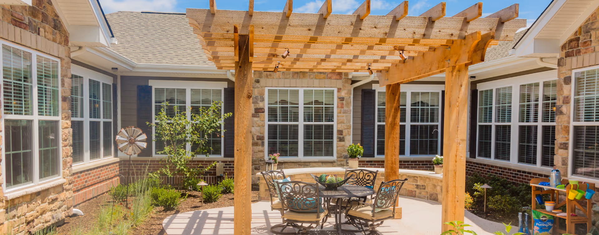 Outdoor courtyard area with a wooden pergola providing partial shade over a round metal table and four cushioned chairs. The courtyard is surrounded by a building with stone and brick walls and multiple windows with white blinds. There are small plants and shrubs along the edges, a small windmill decoration, and a wooden shelving unit with gardening supplies on the right side.