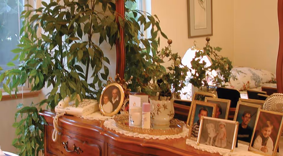 A wooden dresser with a large mirror behind it, decorated with several framed family photos, a potted plant, a telephone, and some personal care items. A bed with floral bedding is partially visible in the background.