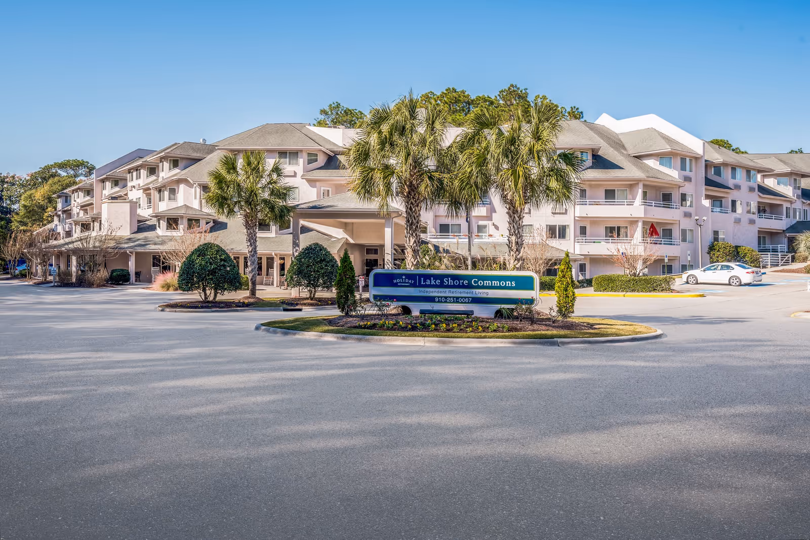 Exterior view of a multi-story senior living facility named Lake Shore Commons with palm trees and landscaped greenery in front, under a clear blue sky.