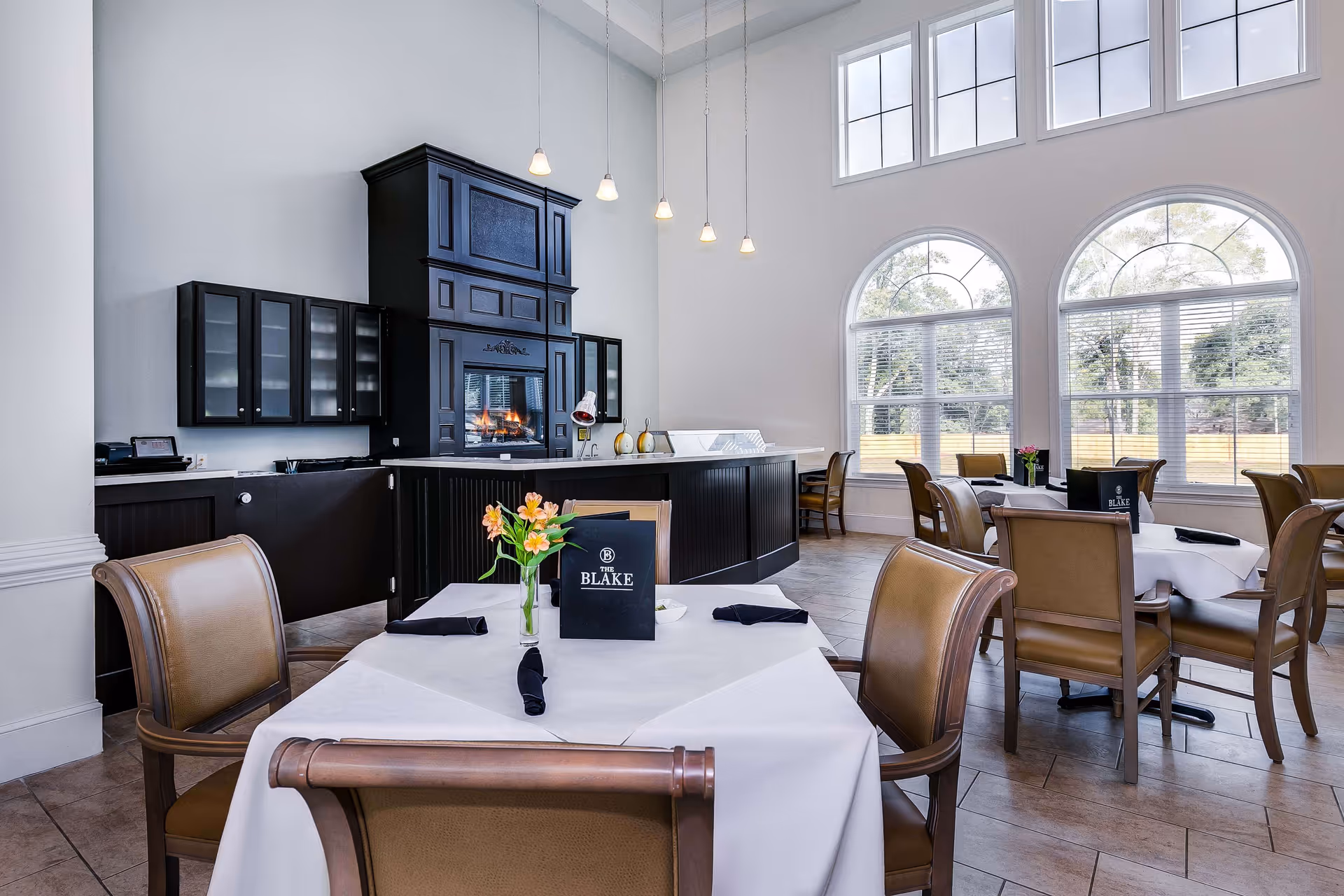 Dining room with tables covered in white tablecloths and set with black napkins, brown chairs, and small flower vases. A black menu with 'The Blake' logo is on the table in the foreground. The room has large arched windows letting in natural light and a black fireplace with a fire burning behind a glass panel. Pendant lights hang from the high ceiling.